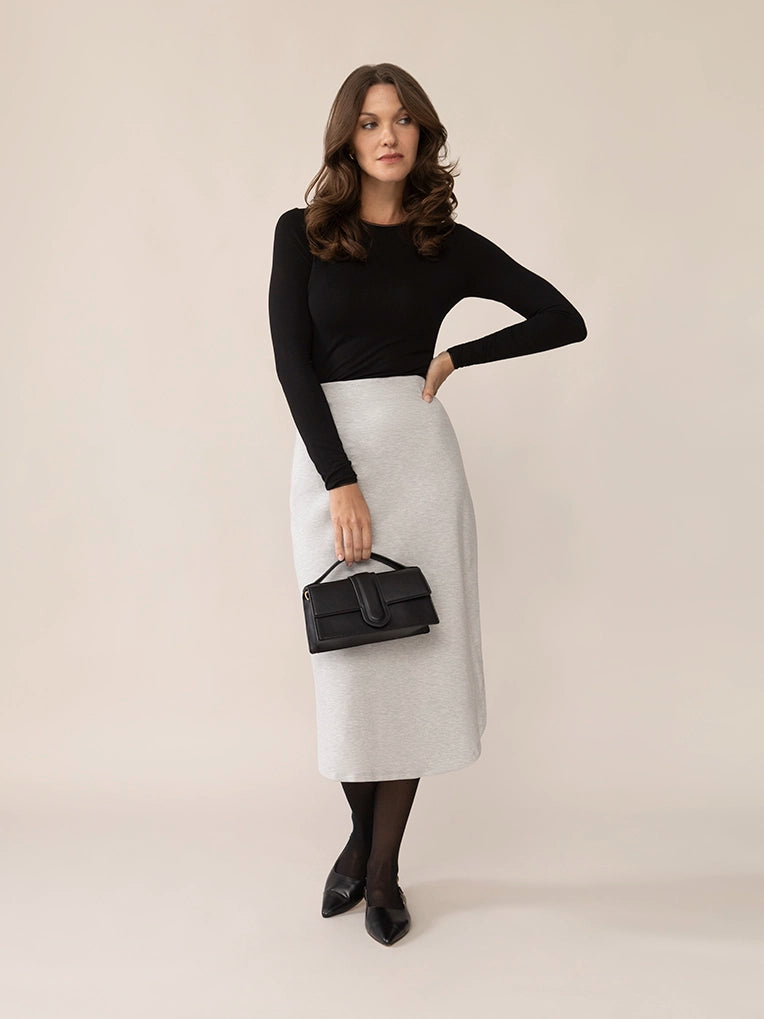 Woman wearing a black top and white skirt holding a black handbag against a plain background