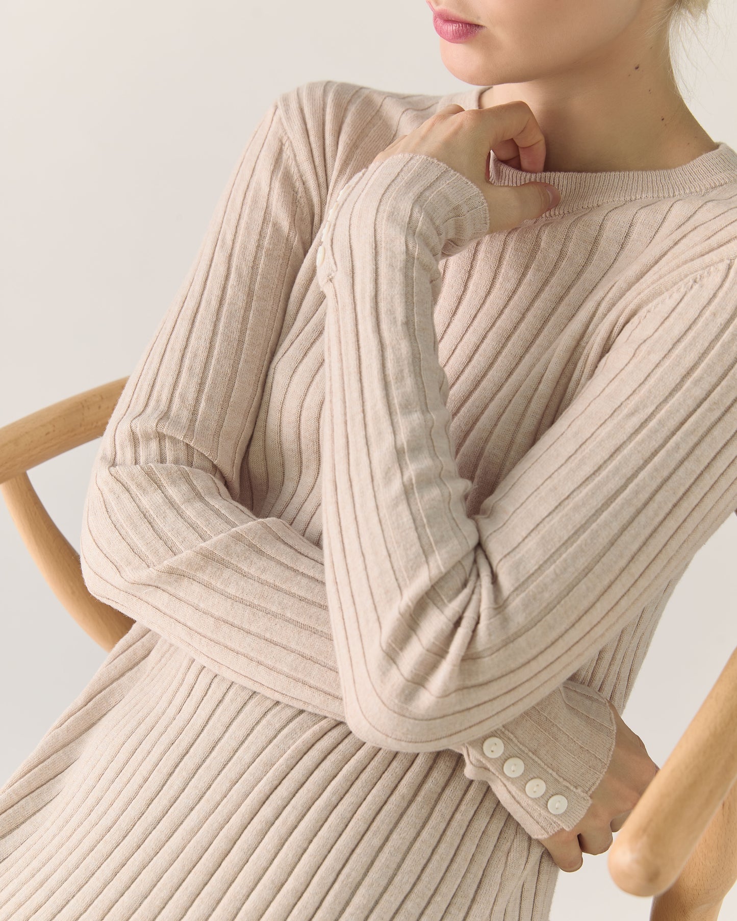 Person wearing a beige ribbed sweater dress with details on sleeve sitting on a wooden chair.