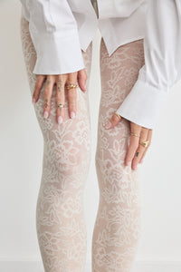 Hands adjusting the Floral Lace tights in Creamy White at the thigh, with a white sleeve cuff visible, showing the floral pattern in the soft cream shade.