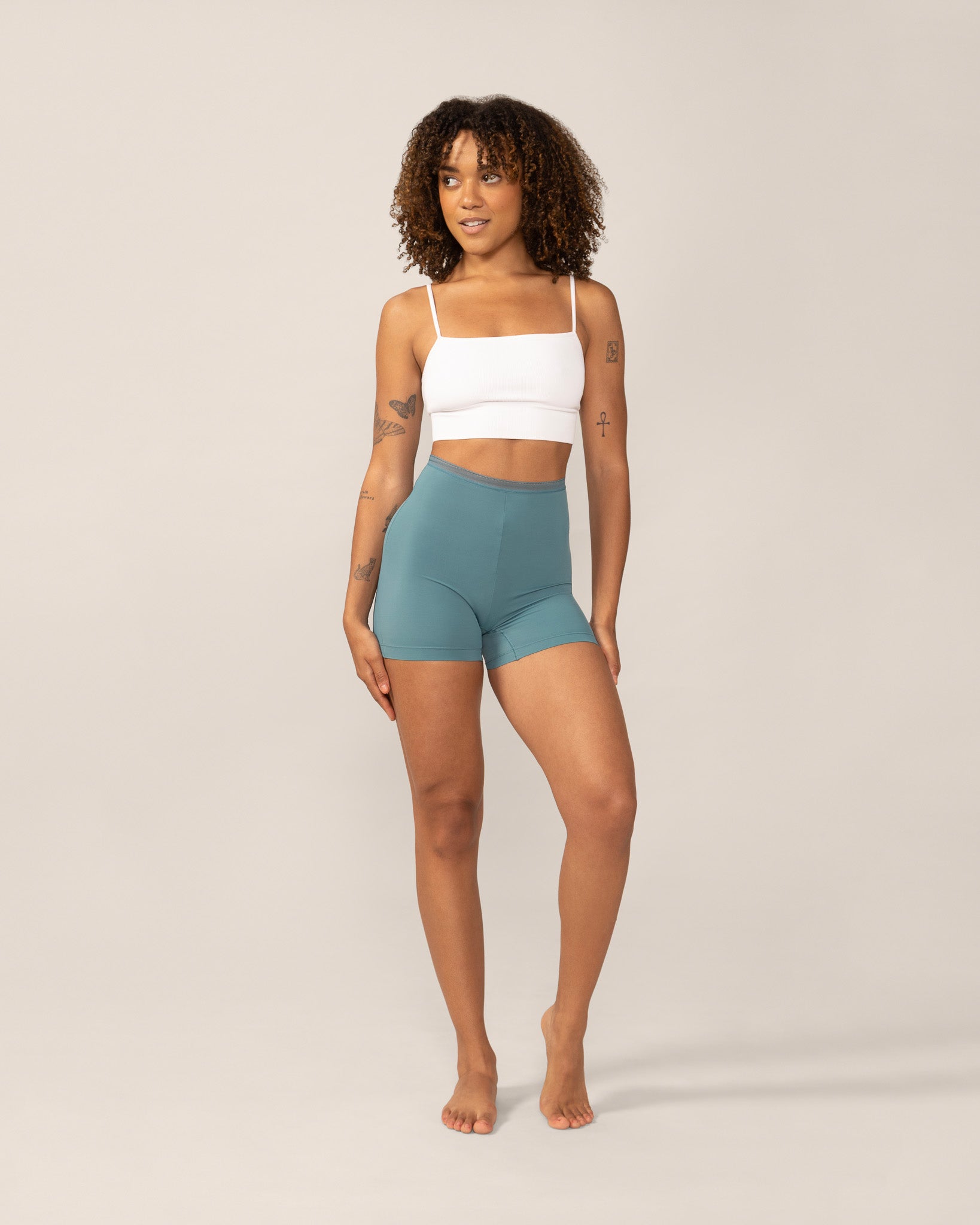 Woman wearing a white sports bra and teal high-waisted anti chaffing shorts on a beige background