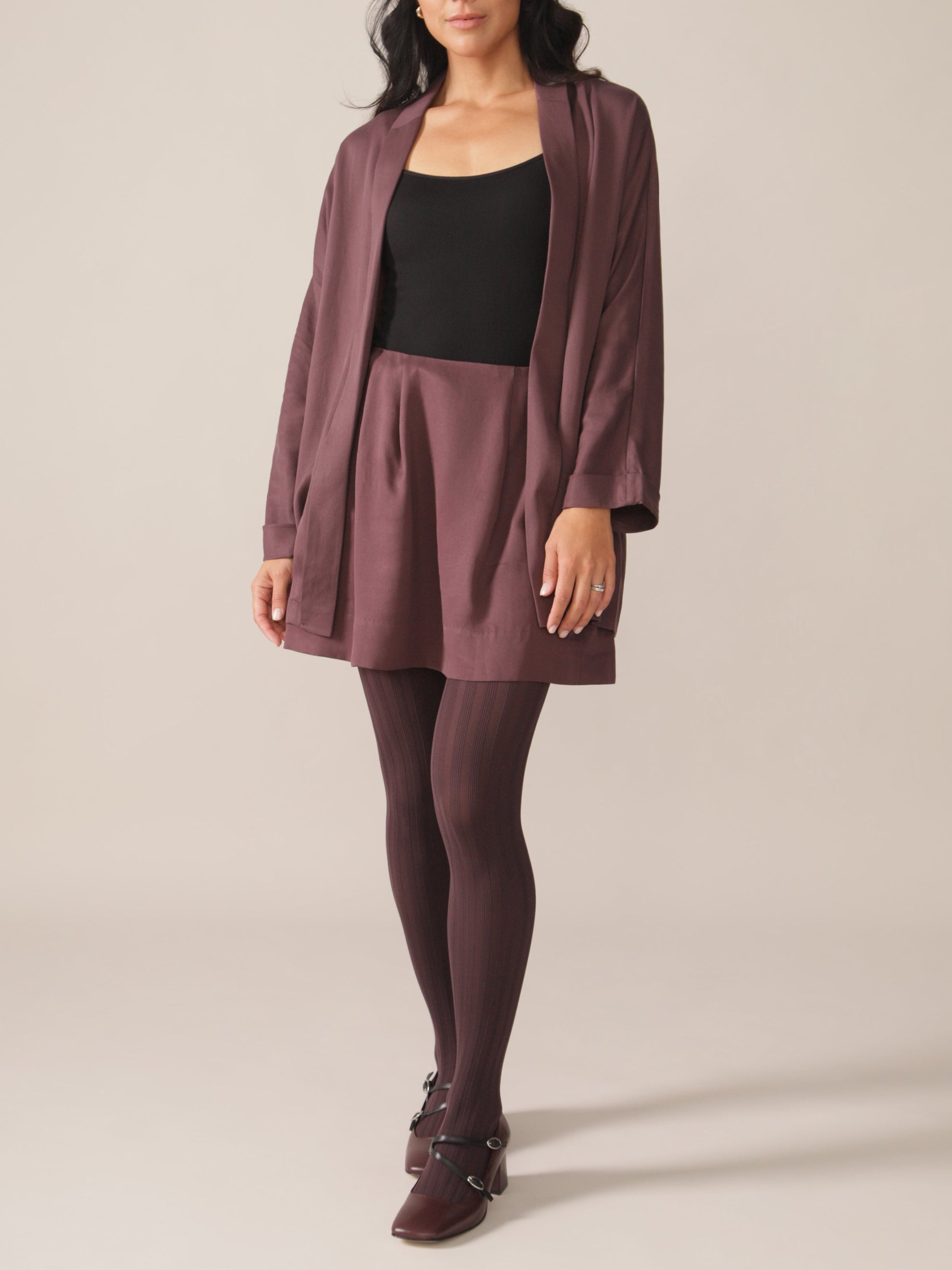 A woman standing wearing a plum purple kimono blazer with a loose fit and a self-tie belt, paired with a plum skirt and tights