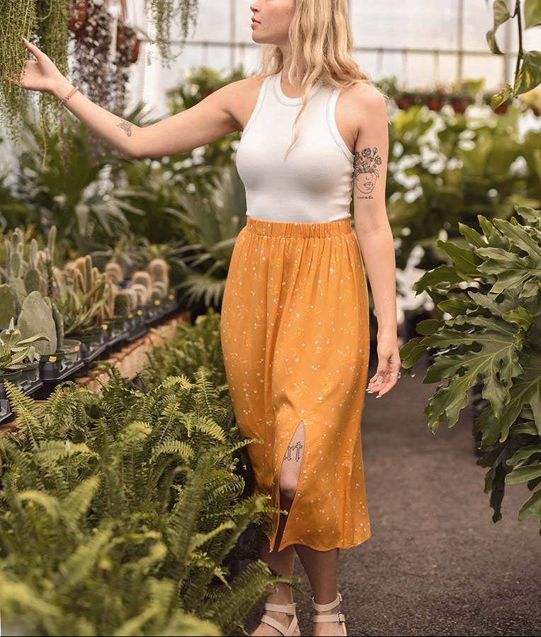 Woman in a white top and yellow skirt standing among plants in a greenhouse.